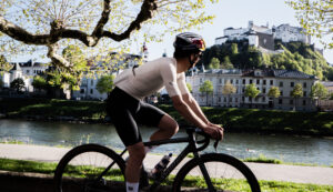 Radfahrer an einem Fluss mit einem Schloss auf einem Hügel und Gebäuden im Hintergrund an einem sonnigen Tag.