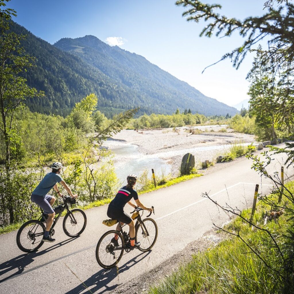 Zwei Radfahrer fahren auf einem gepflasterten Weg durch eine malerische, sonnige Berglandschaft mit Bäumen und einem Fluss in der Nähe.
