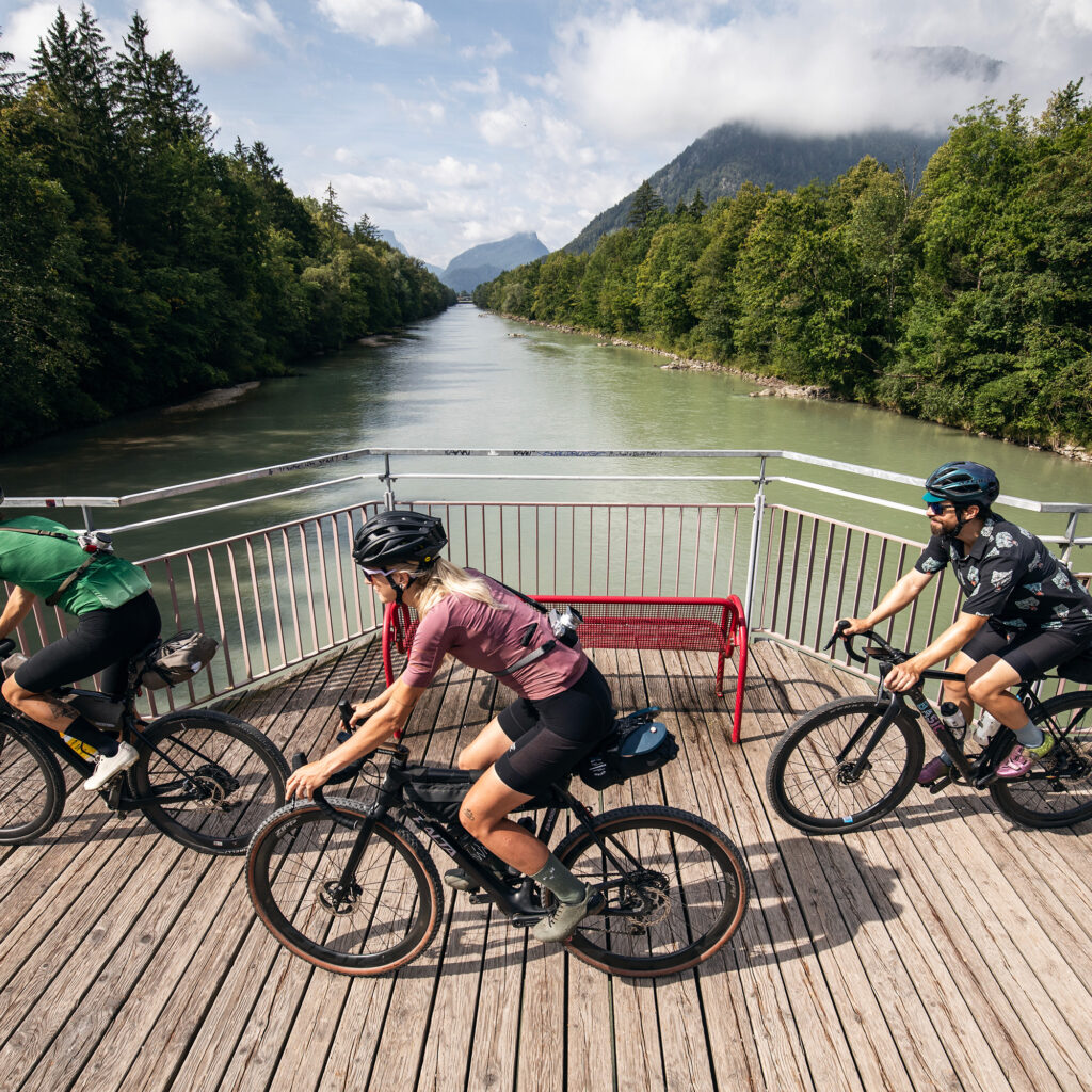 Drei Radfahrer fahren mit ihren Rädern auf einer Holzbrücke über einen Fluss, umgeben von üppigen Bäumen und Bergen.