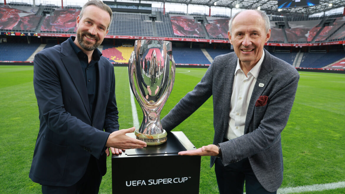 Zwei Männer in Anzügen lächeln, während sie die Trophäe des UEFA-Superpokals auf einem Stadionfeld halten.