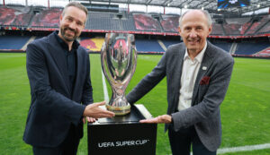 Zwei Männer in Anzügen lächeln, während sie die Trophäe des UEFA-Superpokals auf einem Stadionfeld halten.
