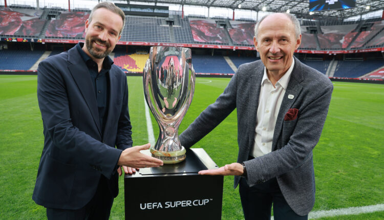 Zwei Männer in Anzügen lächeln, während sie die Trophäe des UEFA-Superpokals auf einem Stadionfeld halten.