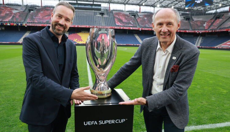 Zwei Männer in Anzügen lächeln, während sie die Trophäe des UEFA-Superpokals auf einem Stadionfeld halten.