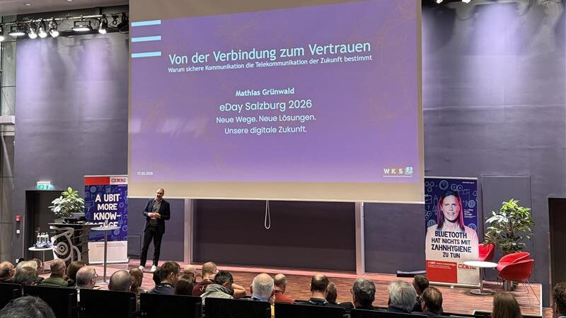 Ein Redner hält einen Vortrag auf der Bühne einer technischen Konferenz, während auf einem Bildschirm der Titel der Veranstaltung angezeigt wird und das Publikum zuschaut.