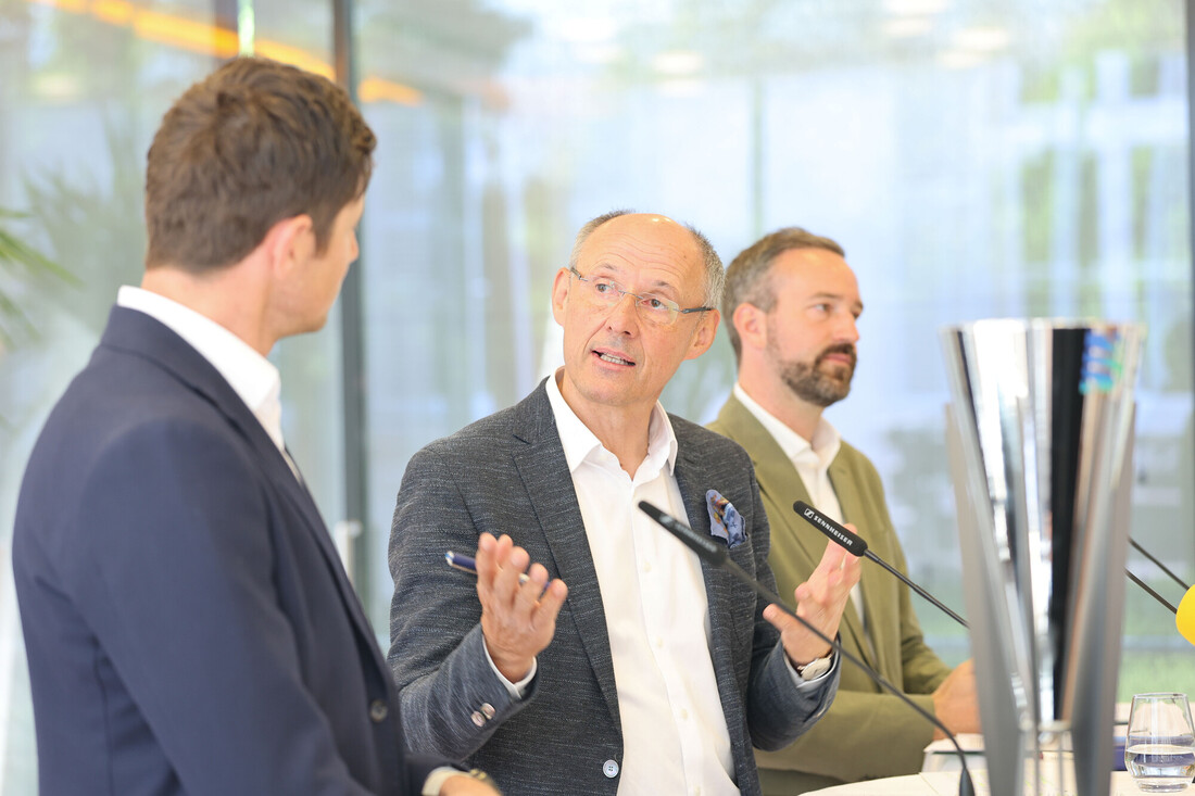 Drei Männer in Anzügen sitzen an einem Tisch und unterhalten sich, im Vordergrund stehen Mikrofone und eine große Trophäe.