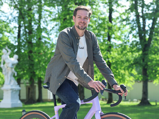 Ein Mann in Freizeitkleidung lächelt, während er auf einem lila Fahrrad in einem sonnigen, baumbestandenen Park sitzt.