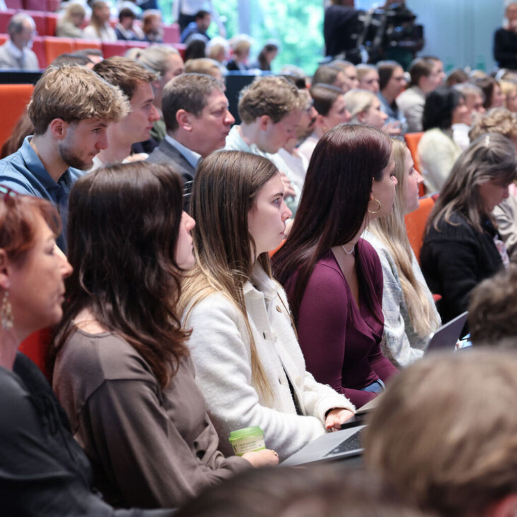 Studierende sitzen in Reihen in einem Hörsaal und hören einem Redner aus dem Off aufmerksam zu.