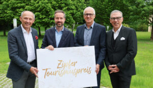 Vier Männer in Anzügen halten ein Schild mit der Aufschrift Zipfer Tourismuspreis im Freien mit grünen Bäumen im Hintergrund.