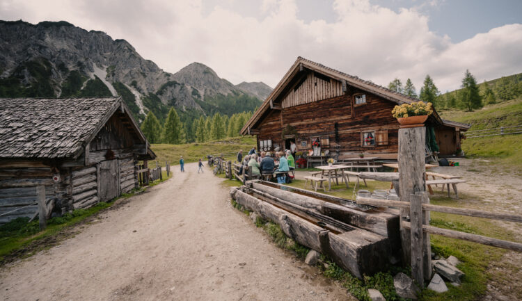 Menschen sitzen an Tischen im Freien bei rustikalen Holzhütten in einem Bergtal mit Bäumen und bewölktem Himmel.