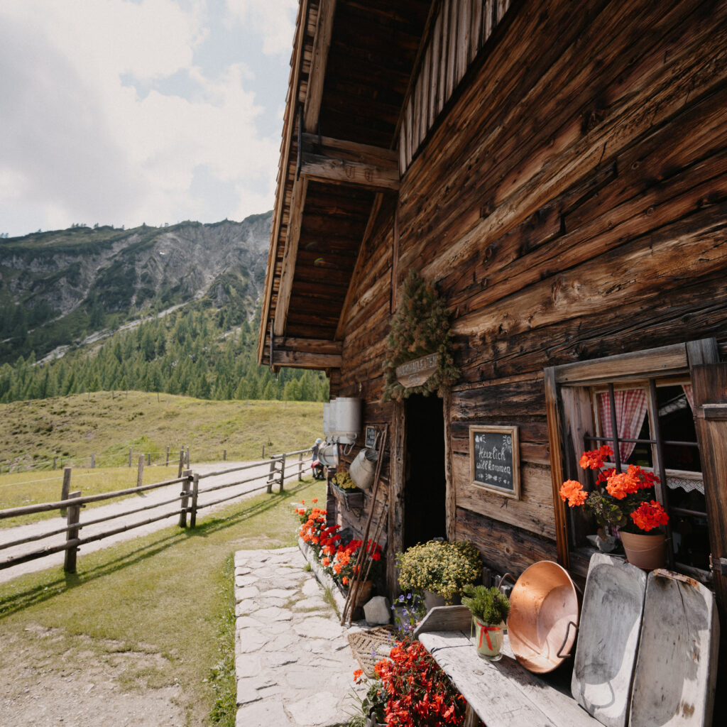 Eine rustikale Holzhütte mit Blumen und Gartengeräten neben einem Zaun und einer grünen Berglandschaft.