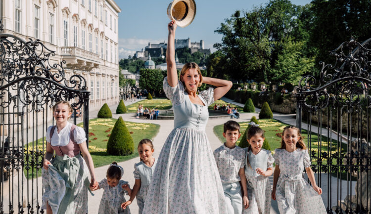 Eine Frau und sechs Kinder in einheitlichen Outfits schlendern an einem sonnigen Tag fröhlich durch kunstvoll verzierte Gartentore.