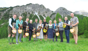 Zwölf Menschen stehen auf einer Wiese vor den Bergen, halten bunte Kuhglocken in den Händen und lächeln in die Kamera.