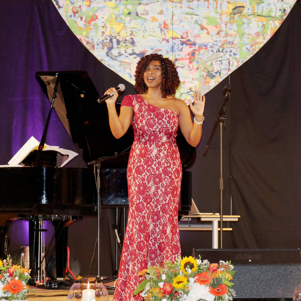 Bei der Veranstaltung „Kulturelle Sommer-Highlights“ singt eine Frau in einem roten Kleid auf der Bühne neben einem Pianisten.