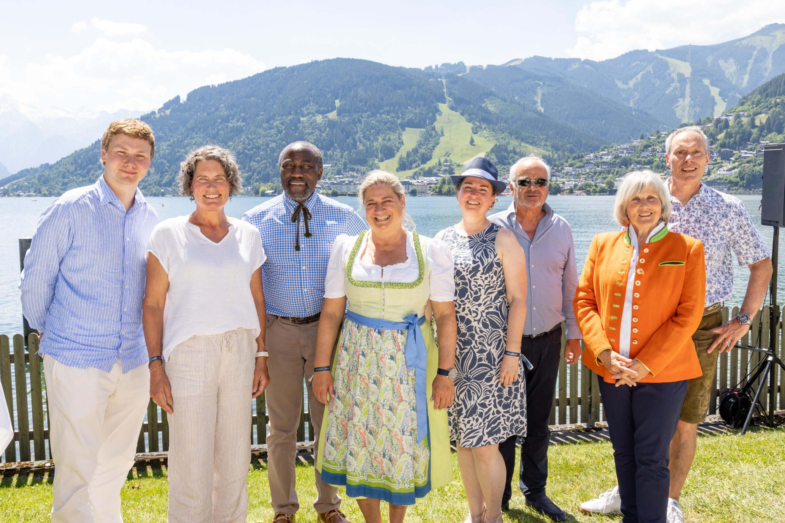 Acht Menschen, die an einem sonnigen Tag im Freien an einem See lächeln, im Hintergrund Berge und grüne Hügel.