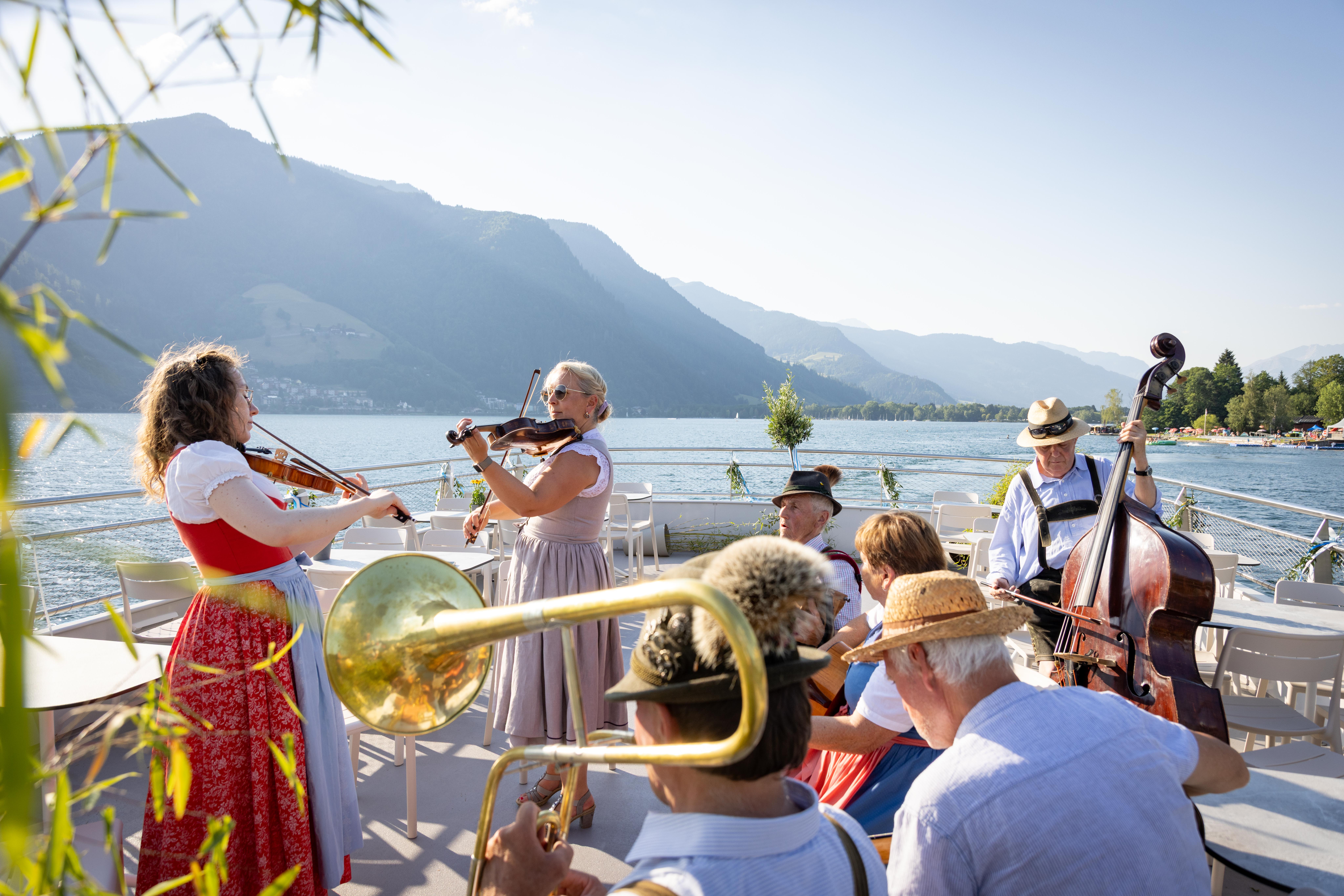 Musiker spielen auf einem Boot Instrumente, im Hintergrund sind Berge und ein See zu sehen, über ihnen erstreckt sich ein klarer Himmel.