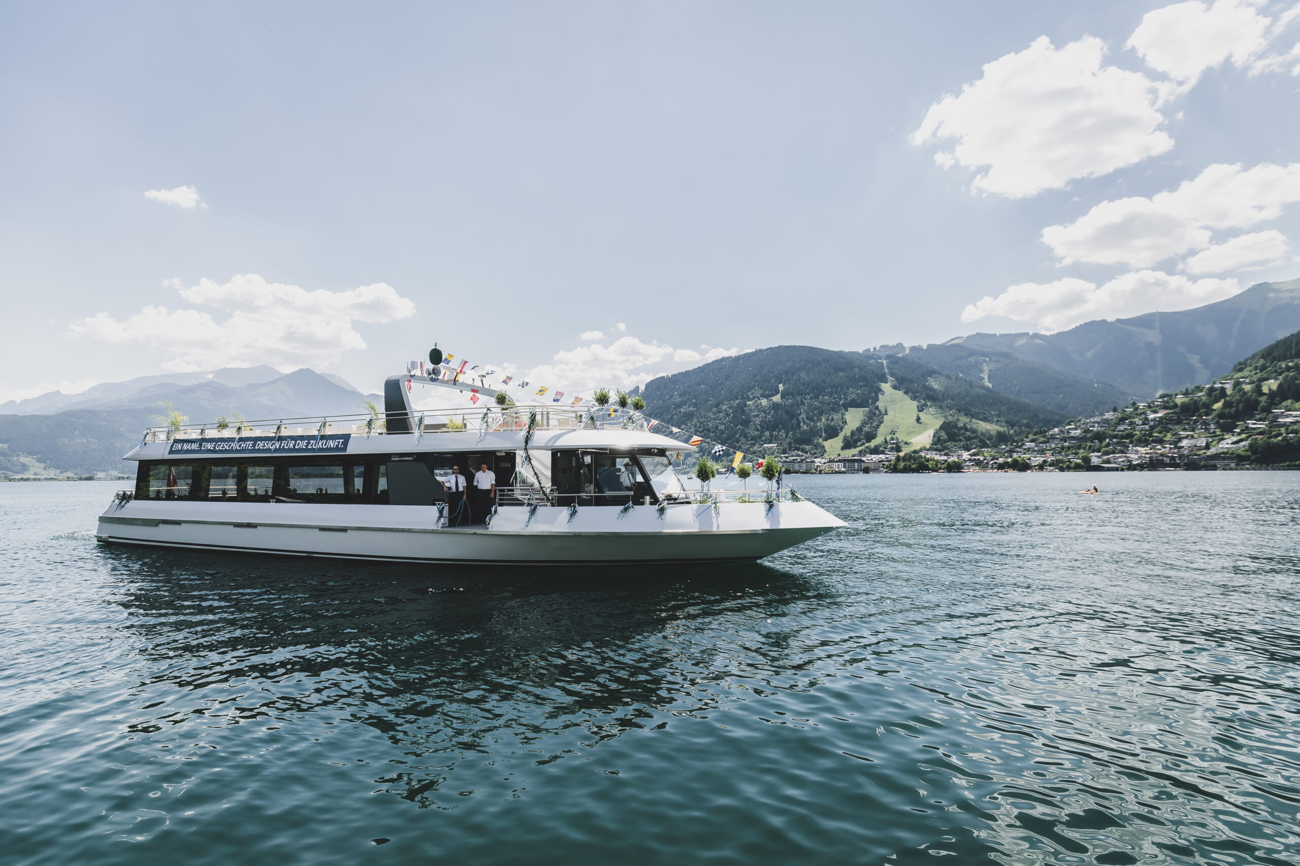 Ein weißes, mit Blumen geschmücktes Ausflugsboot schwimmt auf einem See, im Hintergrund sind Berge und der blaue Himmel zu sehen.