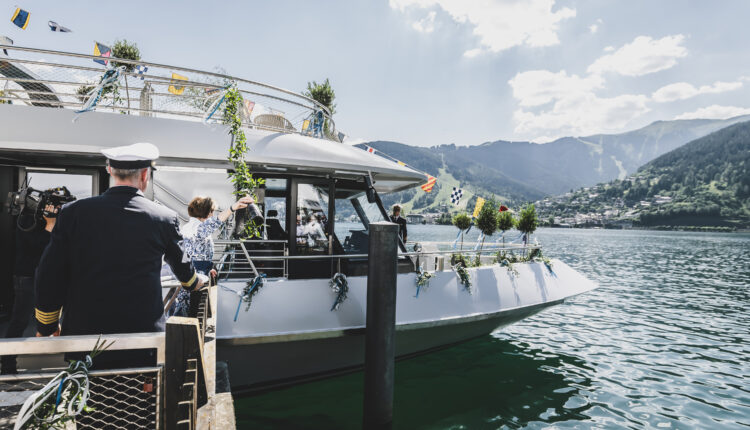 Menschen steigen an einem sonnigen Tag auf einem See, vor dessen Hintergrund sich Berge und eine Stadt abzeichnen, in ein geschmücktes Boot ein.