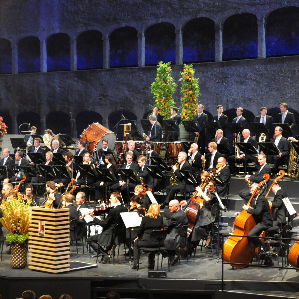 Ein Orchester sorgt für sommerliche Höhepunkte und spielt unter dramatischer Beleuchtung mit Streichern, Blechbläsern und Holzbläsern.