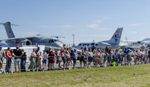 Eine Menschenmenge, die an einem sonnigen Tag bei einer Flugshow im Freien Flugzeuge beobachtet, im Hintergrund stehen Jets.