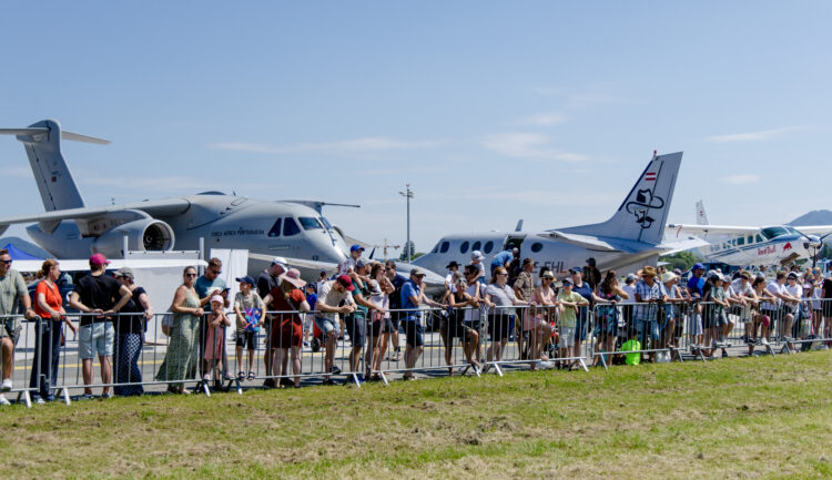 Eine Menschenmenge, die an einem sonnigen Tag bei einer Flugshow im Freien Flugzeuge beobachtet, im Hintergrund stehen Jets.