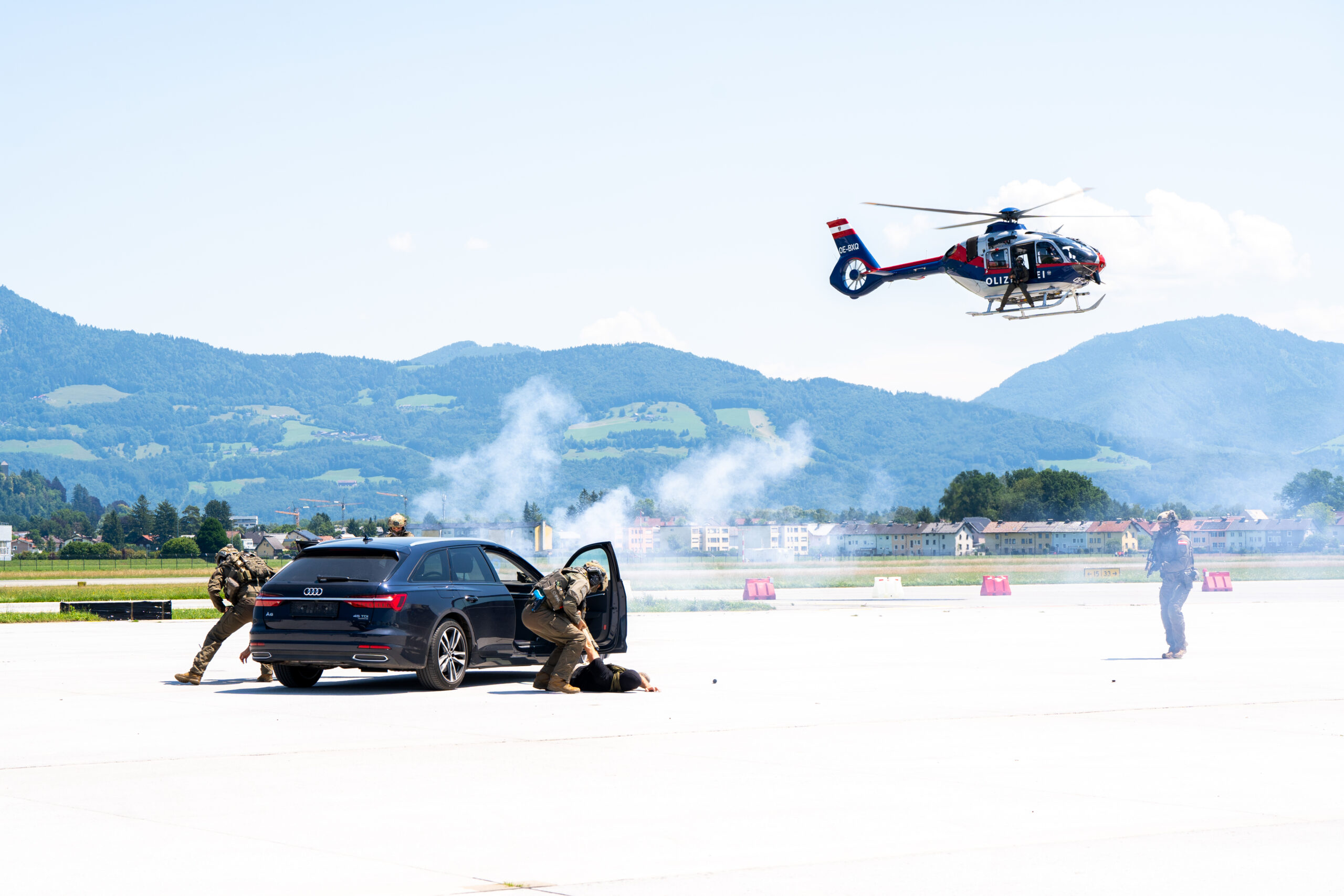 Bewaffnete Soldaten suchen hinter einem Auto Deckung, während ein Hubschrauber in der Nähe auf einem offenen Flugplatz mit Bergen im Hintergrund in der Luft schwebt.