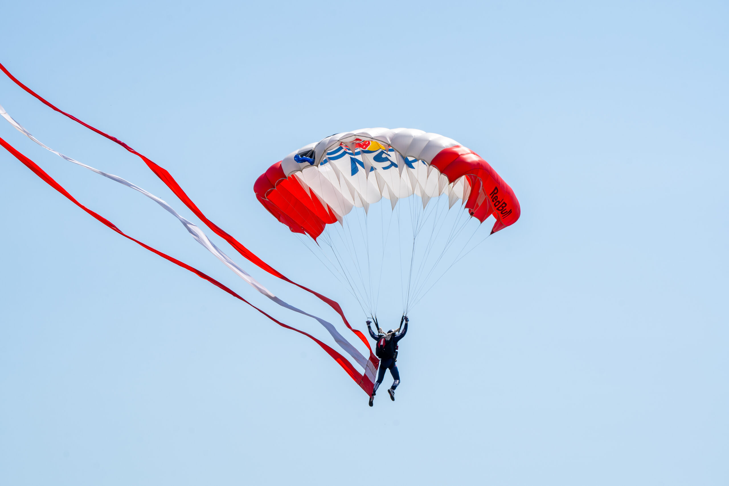 Ein Fallschirmspringer schwebt vor einem strahlend blauen Himmel mit einem Red-Bull-Fallschirm und langen rot-weißen Luftschlangen herab.