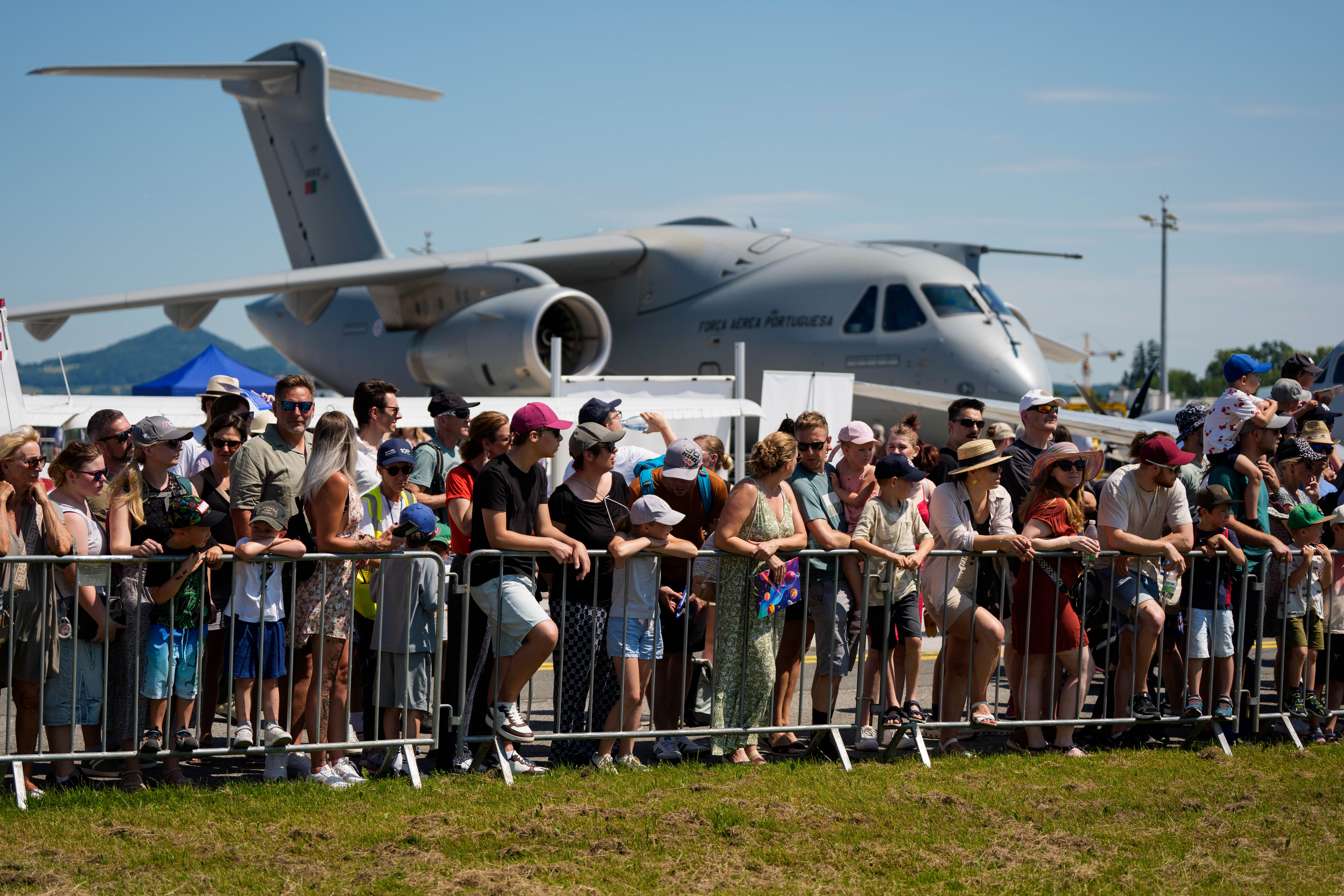 Hinter einer Absperrung steht eine Menschenmenge und verfolgt eine Flugshow, im Hintergrund ist ein großes Militärflugzeug zu sehen.