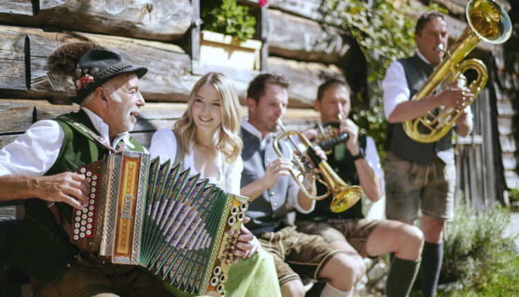 Fünf Menschen in traditioneller österreichischer Tracht spielen im Freien neben einem Holzgebäude auf Instrumenten, lächeln und genießen die Musik.