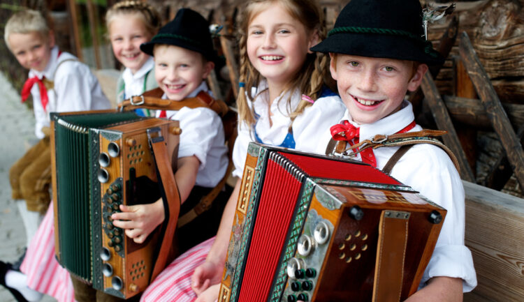 Lächelnde Kinder in traditioneller österreichischer Tracht sitzen auf einer Bank und spielen Akkordeon.