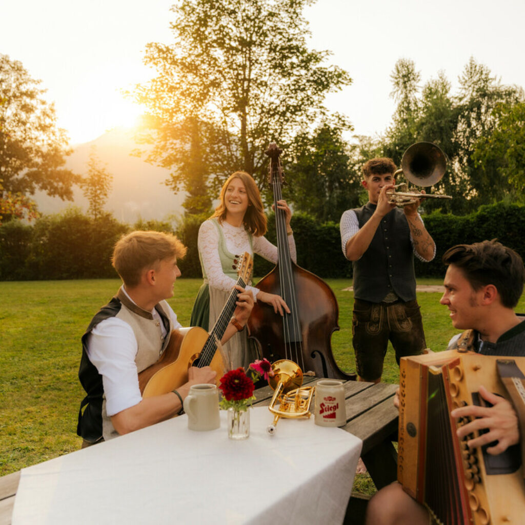 Vier Menschen in traditioneller Tracht spielen im Freien auf Instrumenten – echte kulturelle Sommer-Highlights bei Sonnenuntergang.