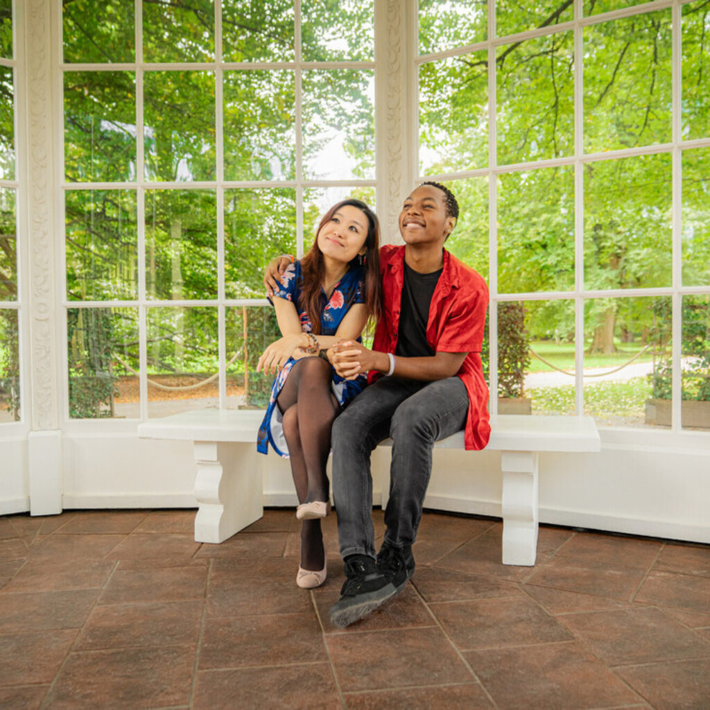 Zwei Menschen sitzen eng beieinander, lächeln und genießen kulturelle Sommer-Highlights in einem Pavillon mit Blick auf den Garten.