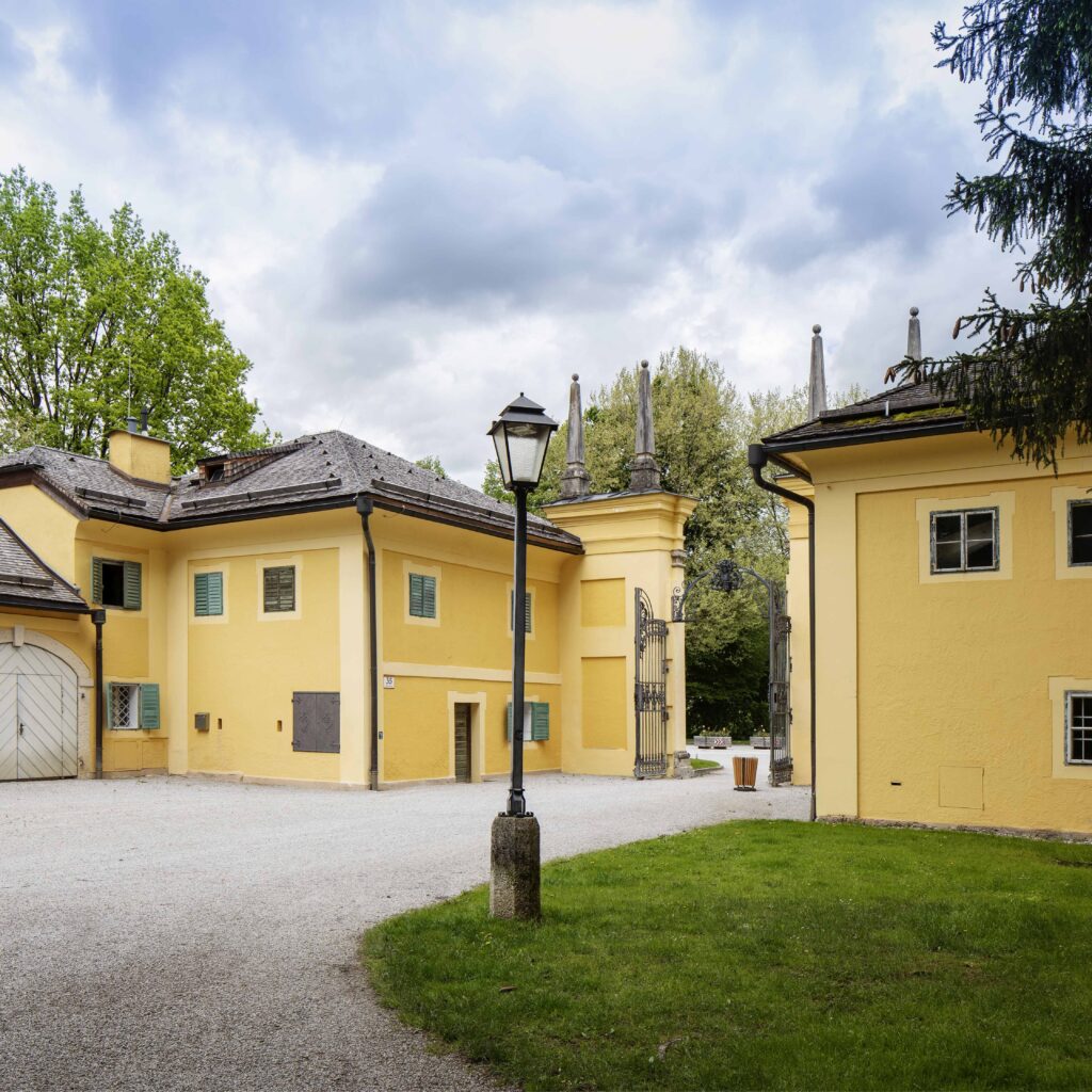 Gelbe Stuckgebäude mit weißen Zierleisten, ein mit Kies bedeckter Innenhof, grünes Gras, Bäume und eine Straßenlaterne unter bewölktem Himmel.