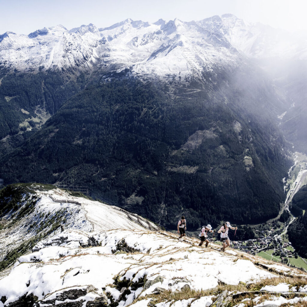 Vier Wanderer steigen einen verschneiten Bergrücken hinauf, im Hintergrund sind ein Tal und schneebedeckte Gipfel zu sehen.