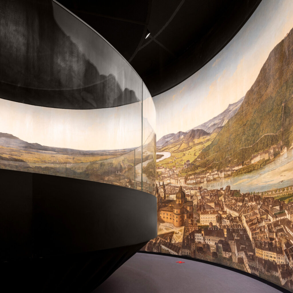 Ein geschwungenes Panorama-Wandbild, das eine Stadt und eine Landschaft zeigt, säumt einen modernen, schwach beleuchteten Ausstellungsraum.