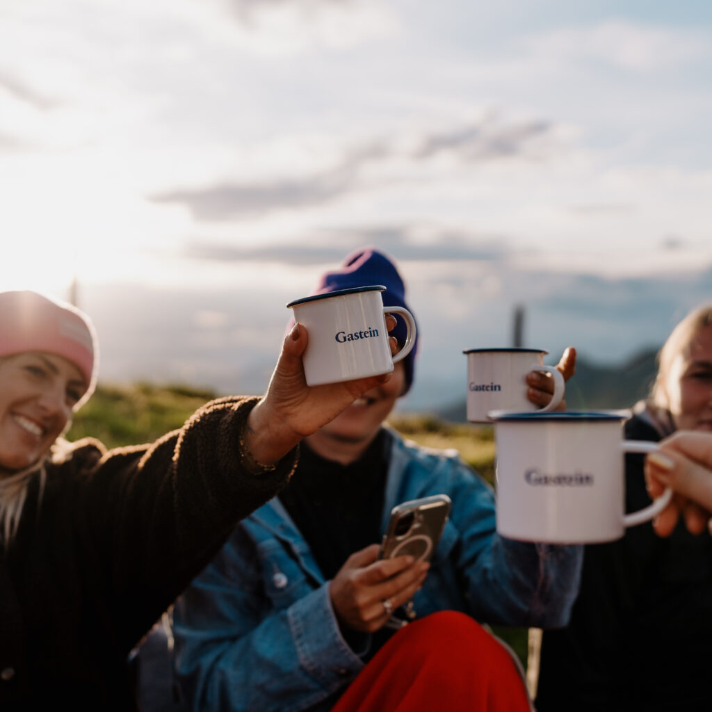 Vier Menschen im Freien stoßen mit weißen Bechern an, lächeln und genießen die Sonne – #SocialMediaSommer2026.