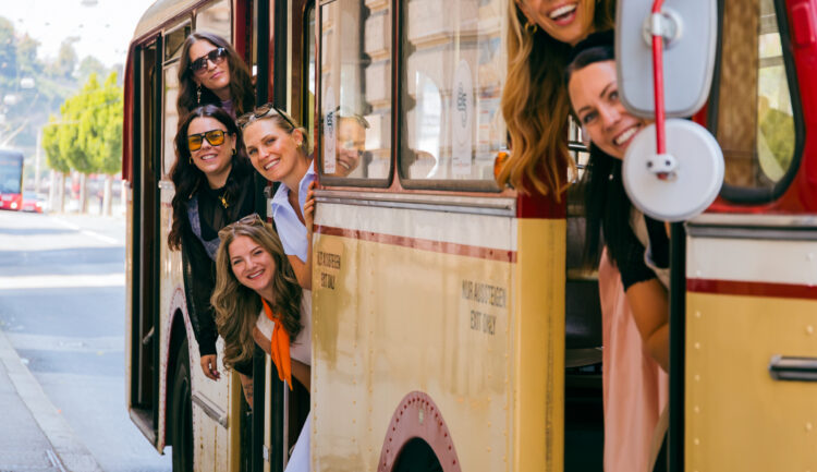Sechs lächelnde Frauen lehnen sich aus einem gelben Oldtimer-Bus und sind bereit für den „Social Media Sommer 2026“ auf einer sonnigen Straße.
