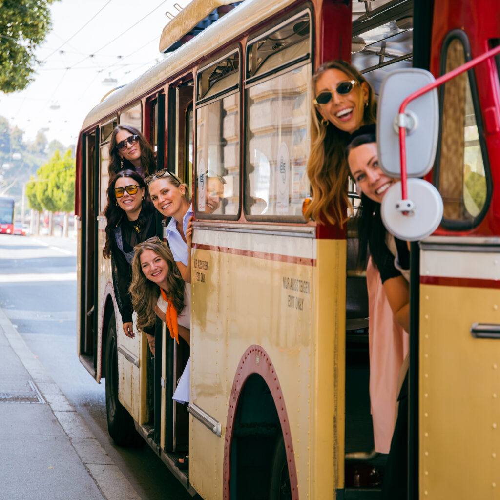 Sechs lächelnde Frauen lehnen sich aus einem alten gelben Bus – Social-Media-Spaß im Sommer 2026 an einem sonnigen Tag.
