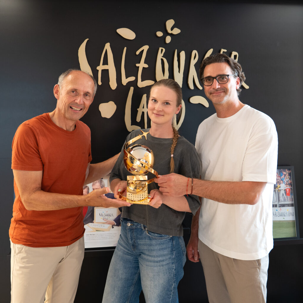 Drei Personen stehen lächelnd vor einem Schild mit der Aufschrift „Salzburger Land“ und halten eine goldene Trophäe in den Händen.