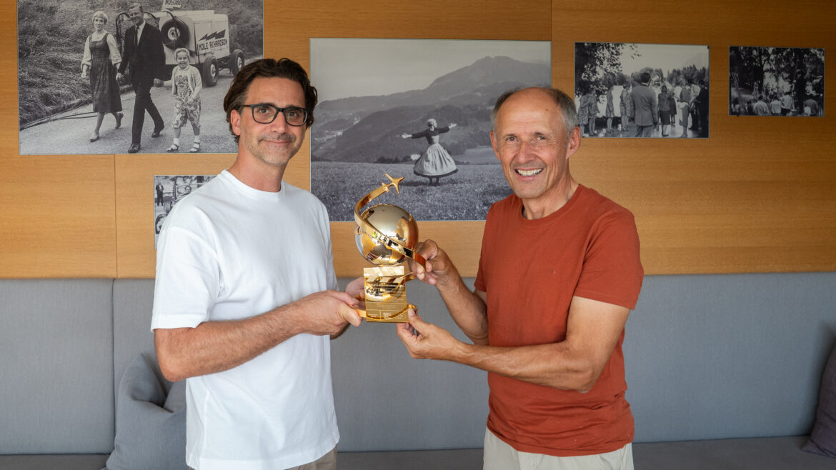 Zwei Männer, die lächeln und gemeinsam eine goldene Trophäe in den Händen halten, in einem Raum, an dessen Wänden Schwarz-Weiß-Fotos hängen.