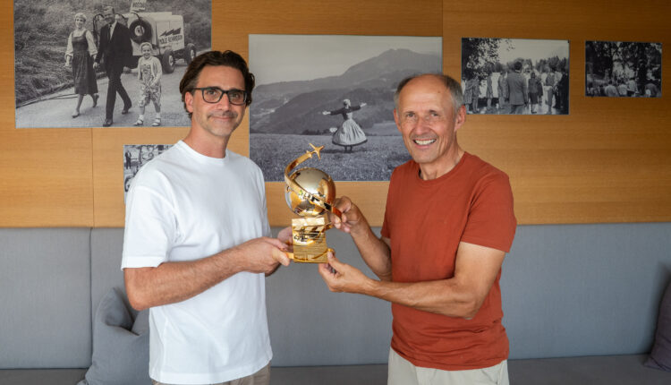 Zwei Männer, die lächeln und gemeinsam eine goldene Trophäe in den Händen halten, in einem Raum, an dessen Wänden Schwarz-Weiß-Fotos hängen.