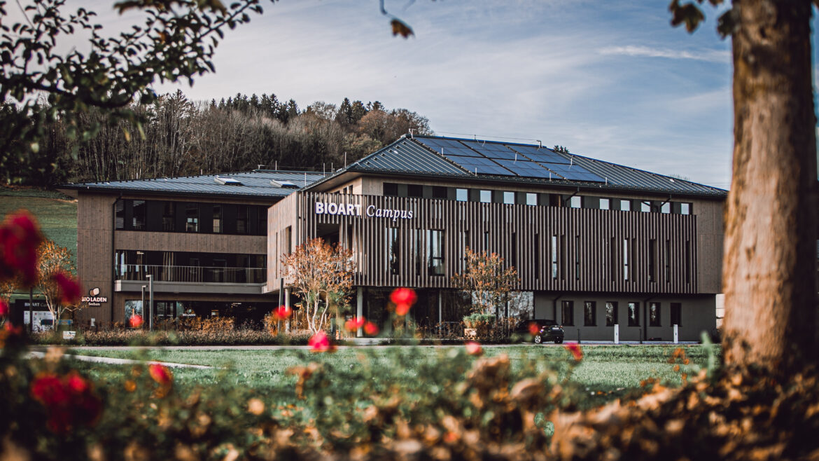 Modernes Gebäude mit der Aufschrift „BIOART Campus“ im Bio-Dorf Seeham, umgeben von Grünflächen, roten Blumen und Bäumen.