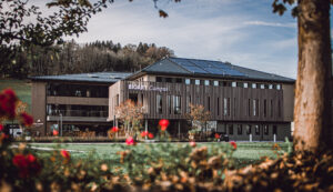 Modernes Gebäude mit der Aufschrift „BIOART Campus“ im Bio-Dorf Seeham, umgeben von Grünflächen, roten Blumen und Bäumen.