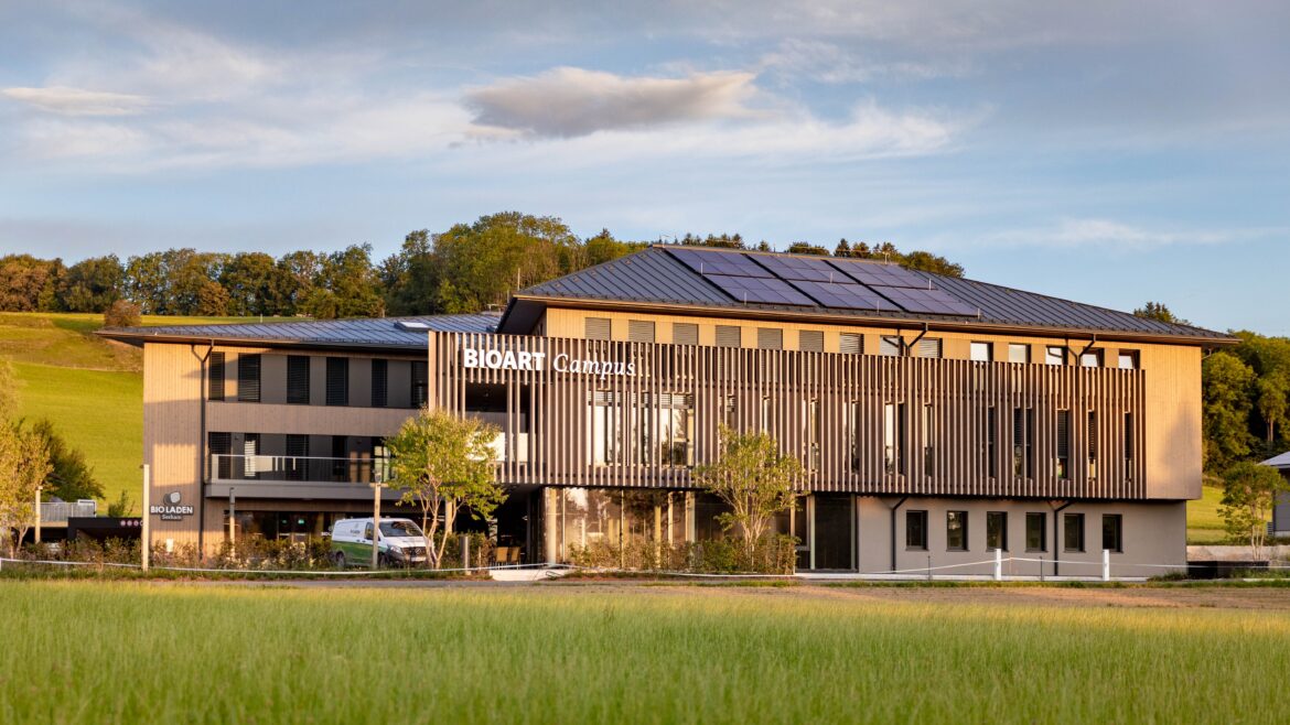 Ein modernes dreistöckiges Gebäude mit der Aufschrift „BioArt Campus“, umgeben von Solarmodulen, Bäumen und grünen Wiesen.