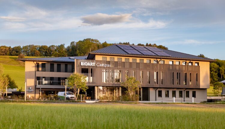 Ein modernes dreistöckiges Gebäude mit der Aufschrift „BioArt Campus“, umgeben von Solarmodulen, Bäumen und grünen Wiesen.