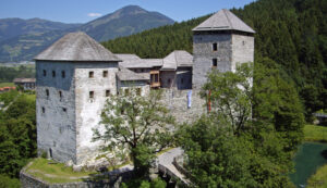 Eine Burg aus Stein mit zwei Türmen, umgeben von Bäumen und Bergen unter einem strahlend blauen Himmel.