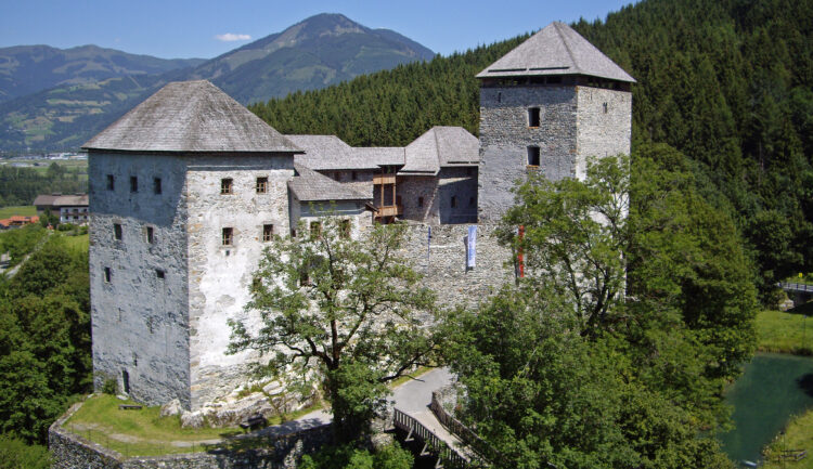 Eine Burg aus Stein mit zwei Türmen, umgeben von Bäumen und Bergen unter einem strahlend blauen Himmel.