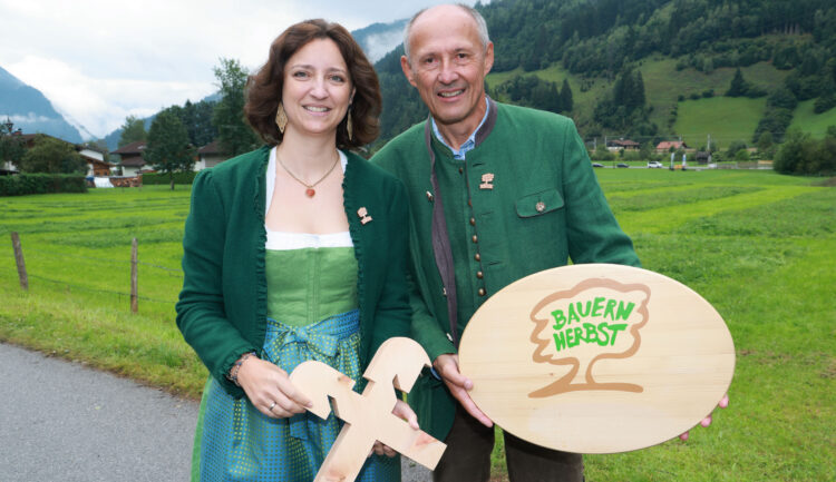 Zwei Personen in traditioneller Tracht halten im Freien Schilder mit der Aufschrift „Bauernherbst-Kooperation mit Raiffeisen“ und einer Baumgrafik in der Hand.