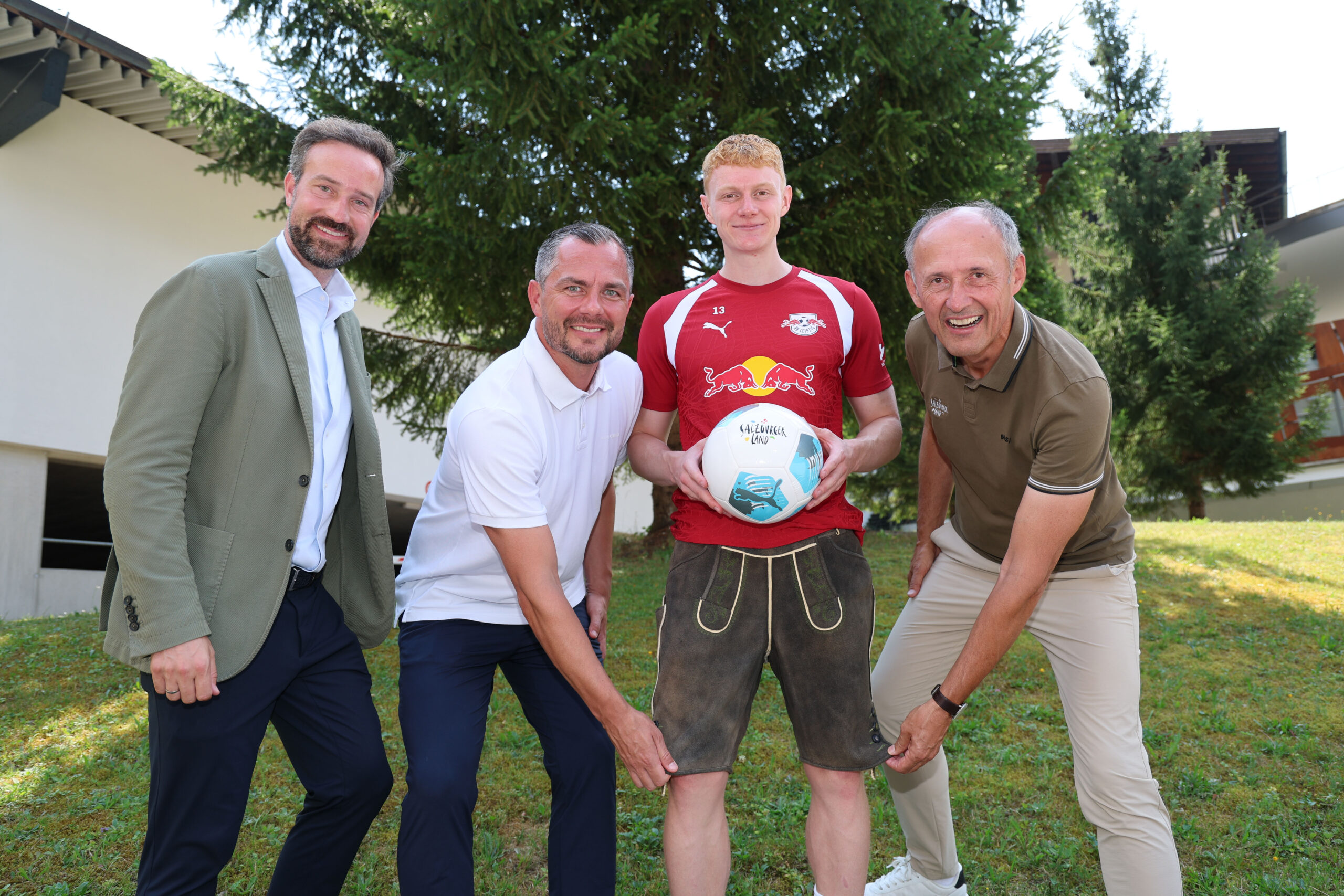 Vier Männer posieren im Freien; einer davon in Sportkleidung hält einen Fußball in der Hand
