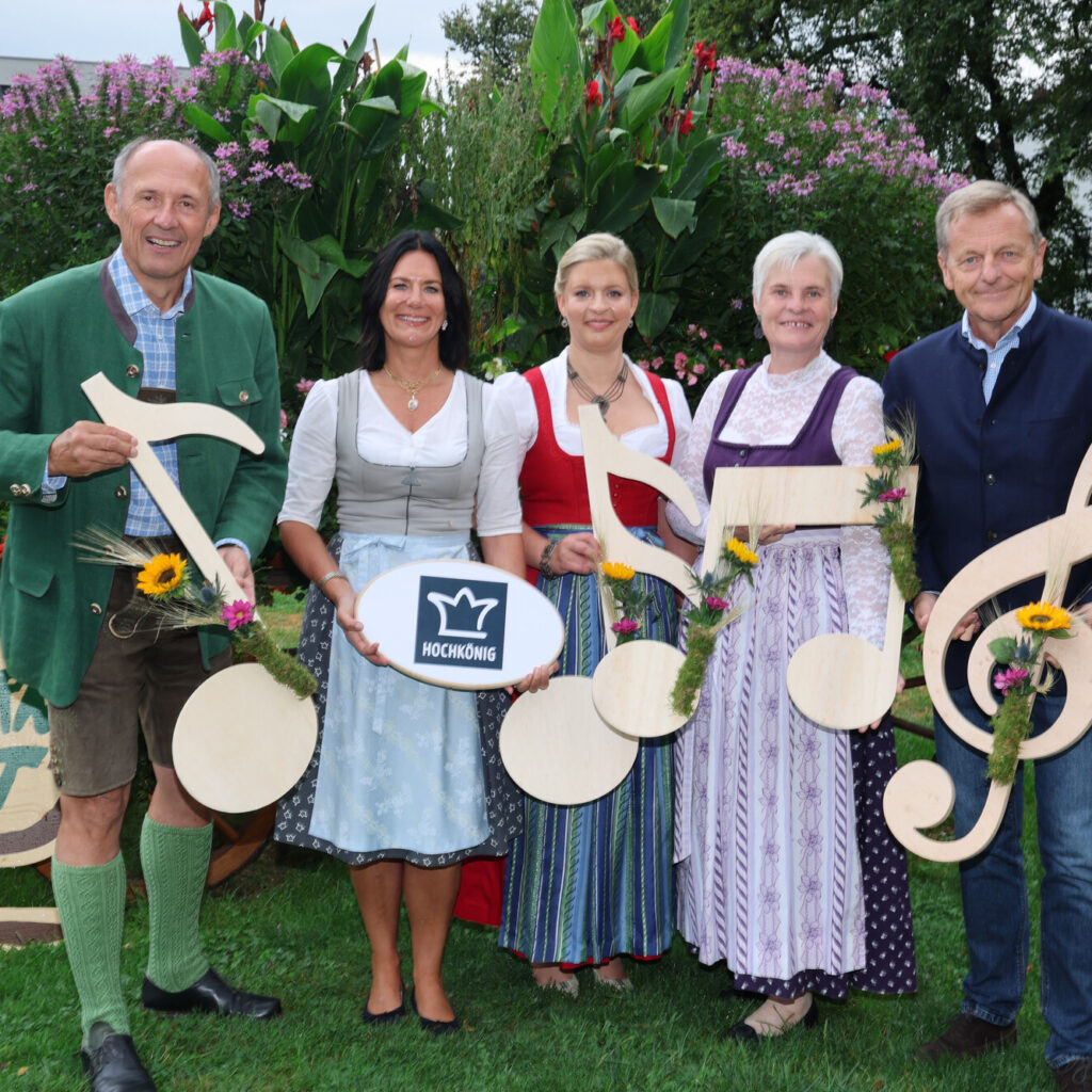 Fünf Personen in traditioneller Tracht halten Noten und Blumen in den Händen und stehen auf einer Wiese neben einem Schild mit der Aufschrift „Bauernherbst“.