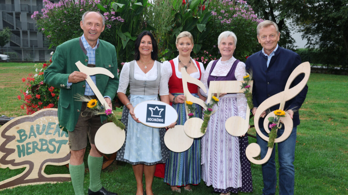 Fünf Personen in traditioneller Tracht halten Noten und Blumen in den Händen und stehen auf einer Wiese neben einem Schild mit der Aufschrift „Bauernherbst“.